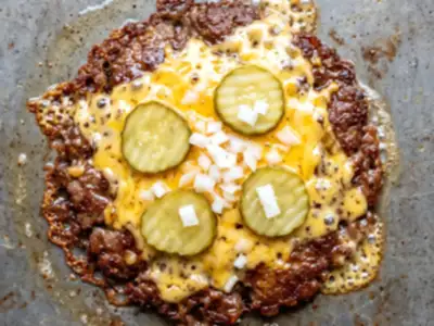 Concept imagery: a smash patty with a browned lacy crust under bubbling melted cheese, pickles and diced onion, on the griddle